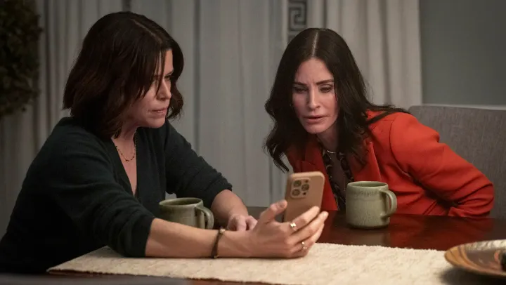 Two brunette women sit at a table with mugs in front of them. They are staring at a phone with concerned expressions.